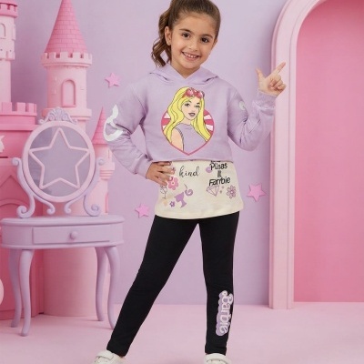 Menina com hoodie lilás, calças pretas com texto Barbie e ténis brancos em cenário rosa com castelo e cadeira decorativa.
