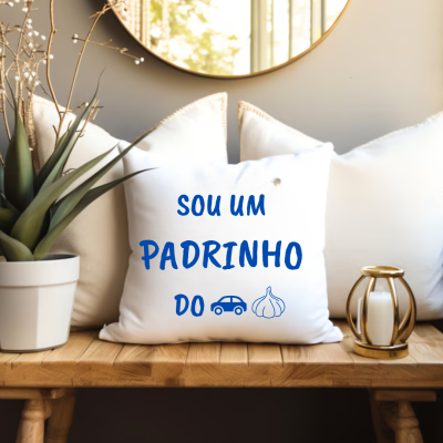 Almofada branca com texto azul 'SOU UM PADRINHO DO' e ícones de carro e alho num ambiente com planta e castiçal