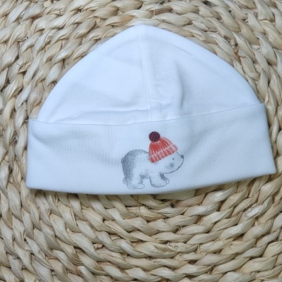Gorro branco para bebé com desenho de urso e gorro vermelho sobre superfície trançada