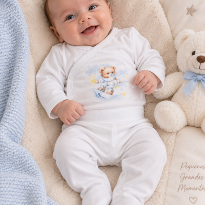 Bebé sorridente vestido com macacão branco com ursinho estampado, deitado em manta bege com cobertor azul e ursinho de peluche ao lado.