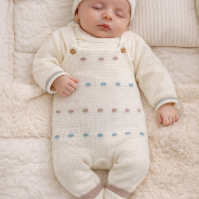Bebé vestido com macacão de malha branco com pontos azuis e castanhos, gorro branco e deitado em cama com cobertores felpudos