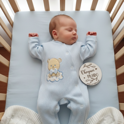 Bebé dormindo roupa azul com ursinho e manta branca na cama com grades de madeira