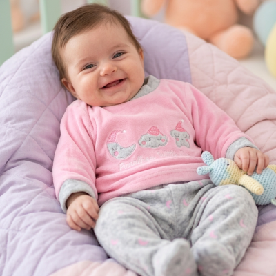 Bebé sorridente vestido com camisola rosa e calças cinzentas, a segurar brinquedo de malha colorido