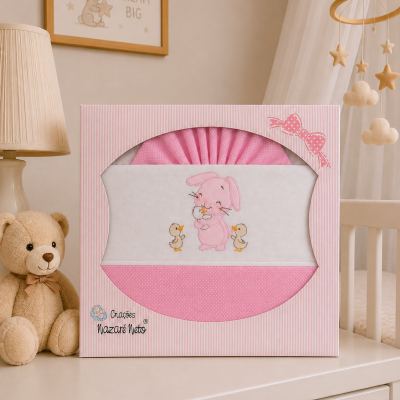 Roupa de cama rosa para bebé embalada em caixa decorada com coelhinho e patos, ao lado urso de peluche castanho.
