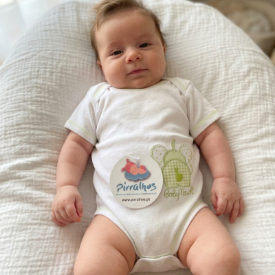 Bebé deitado com body branco com costuras verdes claras e estampa de lagarta verde bordada, etiqueta redonda com logo Pirralhos