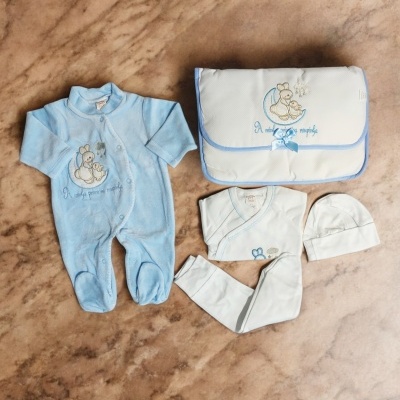 Conjunto de roupa de bebé azul e branco com bolsa, macacão, body, camisola e gorro sobre superfície marrom.