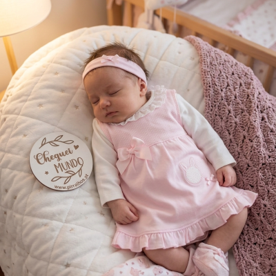 Bebé vestida de rosa a dormir numa almofada branca com manta rosa e berço ao fundo