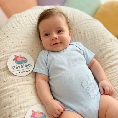 Bebé de body azul claro deitado numa almofada creme com estrelas douradas junto a cartões redondos da marca Pirralhos