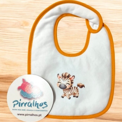 Babete branco com borda laranja e estampa de zebra, sobre superfícies de madeira clara, com cartão da marca Pirralhos.