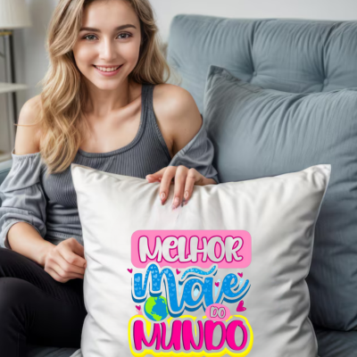 Almofada branca com texto colorido MELHOR MÃE DO MUNDO segurada por mulher num sofá cinza