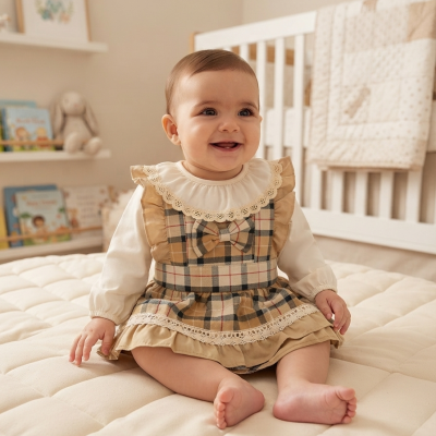 Bebé com vestido xadrez castanho e creme sentado numa cama bege clara em quarto de bebé com berço branco
