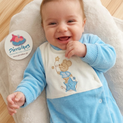 Bebé com macacão azul claro e branco deitado em almofada branca com etiqueta Pirralhos ao lado