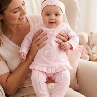 Bebé com conjunto rosa e faixa de cabeça, segurada pela mãe numa poltrona bege claro.