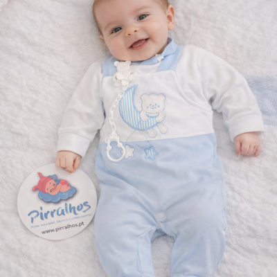 Bebé com macacão azul claro e branco deitado numa manta branca a segurar cartão com o texto Pirralhos.