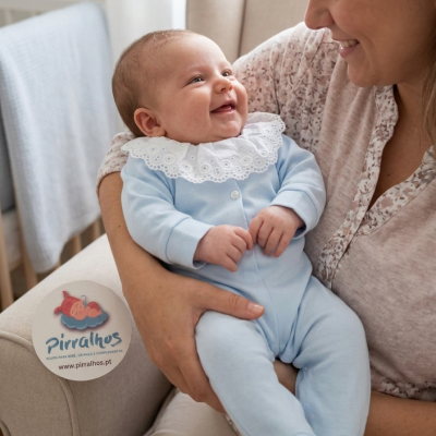 Bebé com macacão azul claro e gola branca de renda, sorrindo enquanto é segurado por mulher num sofá bege.