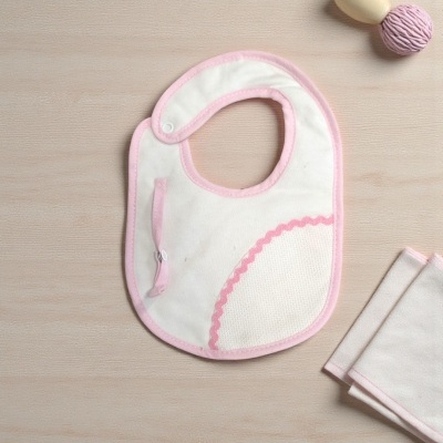 Babete de bebé branco com bordas rosa e bolso, pano e acessório rosa em fundo de madeira clara