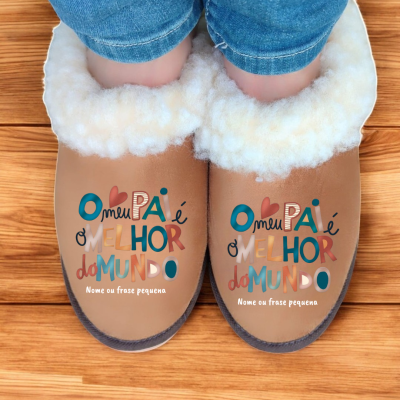 Pantufas castanhas forradas a lã branca com texto colorido no topo, usadas com jeans, em chão de madeira