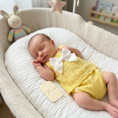 Bebé a dormir com roupão amarelo e laço branco num berço acolchoado com móbil colorido