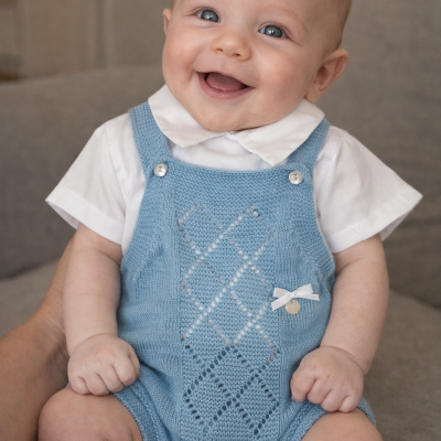 Bebé sorridente com jardineira azul de malha e body branco sentado em sofá cinzento
