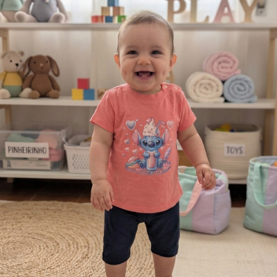 Bebé a sorrir com camisola rosa estampada e calções pretos em quarto de brincar com brinquedos e mantas