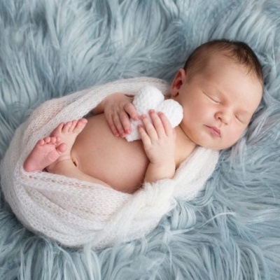 Bebé recém-nascido a dormir enrolado numa manta branca segurando coração de peluche sobre tapete azul
