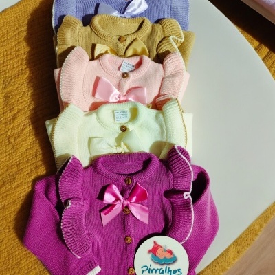 Cinco casacos de malha para bebés coloridos com laços e botões de madeira sobre superfície branca e amarela