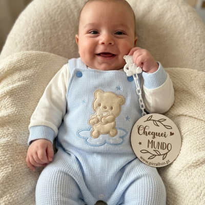 Bebé sorridente vestido com macacão azul claro e branco com ursinho bordado sentado em tecido creme macio segurando plaquinha redonda de madeira.