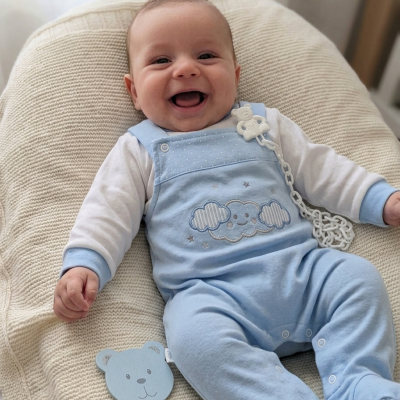 Bebé sorridente com roupa azul e branca sentado em tecido branco de malha com brinquedo de cabeça de urso azul ao lado.