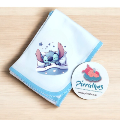 Fralda de bebé branca com borda azul e personagem Stitch a dormir, em mesa de madeira clara, com etiqueta branca redonda ao lado.