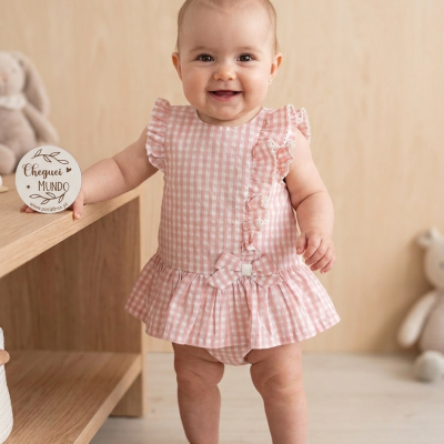 Bebé vestindo roupa rosa xadrez com folhos e laço, segurando cartão redondo com texto em ambiente interior