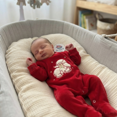 Bebé a dormir de fato vermelho com ovelha branca num berço confortável cinzento e almofada creme