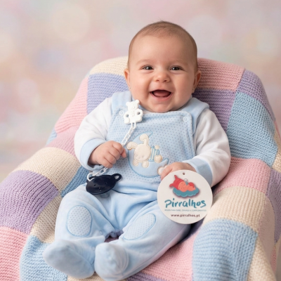 Bebé sorridente com pijama azul e chupeta sentado em poltrona com manta colorida