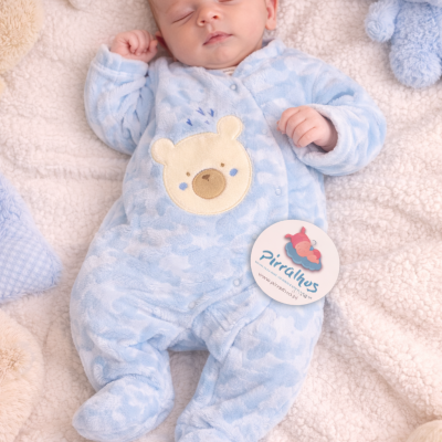 Bebé a dormir vestido com pijama azul claro com urso e etiqueta Pirralhos sobre mantas e almofadas.