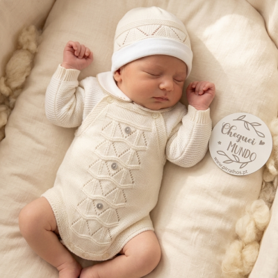 Bebé a dormir vestido de malha branca com gorro, etiqueta com texto Cheguei MUNDO