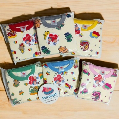 Conjuntos de roupa de bebé com estampados coloridos e golas de cores diferentes, embalados em plástico, sobre madeira clara