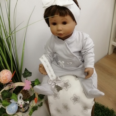 Boneca bebé com pijama cinzento e faixa com laço branca sentada em manta com estrelas num ambiente decorado
