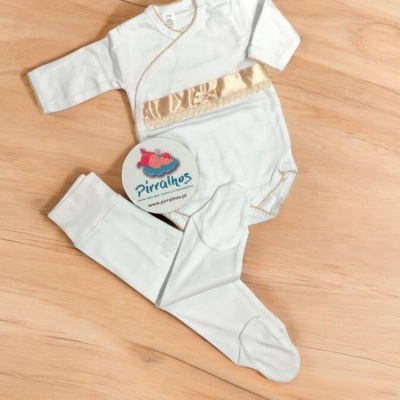 Conjunto de roupa para bebé branco com faixa dourada e calças com pés num chão de madeira clara