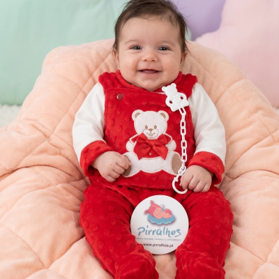 Bebé com pijama vermelho e ursinho sentado numa almofada rosa