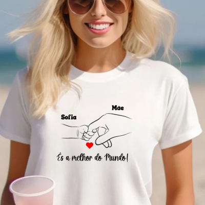Mulher loira de óculos escuros a usar t-shirt branca com design de mãos e texto em português, segurando copo cor-de-rosa na praia.