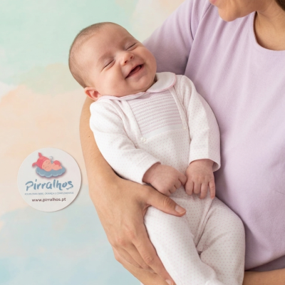 Bebé a sorrir vestido com fato branco de pequenos pontos rosa nos braços de adulto com camisola lilás.