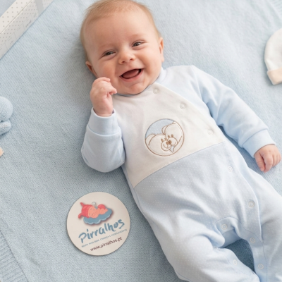 Bebé sorridente vestido com macacão azul e branco sobre manta azul, etiqueta redonda Pirralhos ao lado.