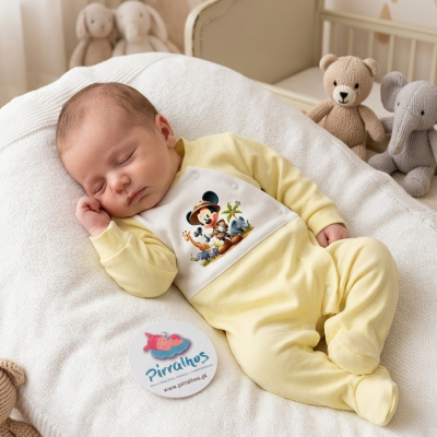 Bebé a dormir com macacão amarelo claro e estampa Mickey Mouse sobre almofada branca tricotada com brinquedos tricotados ao fundo
