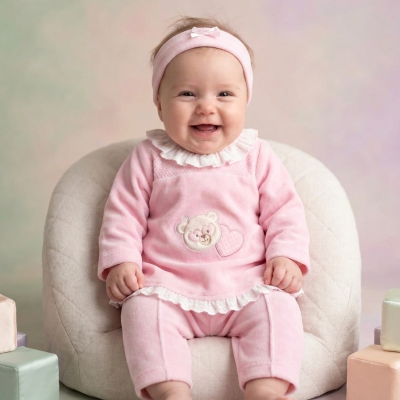 Bebé a sorrir com pijama rosa e faixa de cabeça sentada em cadeira acolchoada creme com blocos coloridos ao lado.