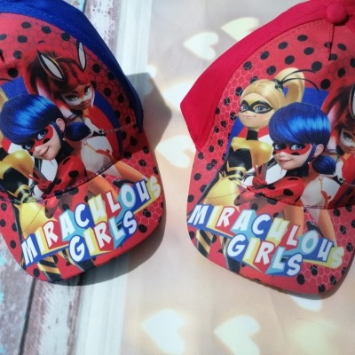 Dois bonés vermelhos e azul com estampas das personagem Miraculous Girls e texto colorido na aba