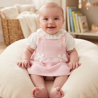 Bebé com vestido rosa e camisa branca sentado numa almofada creme