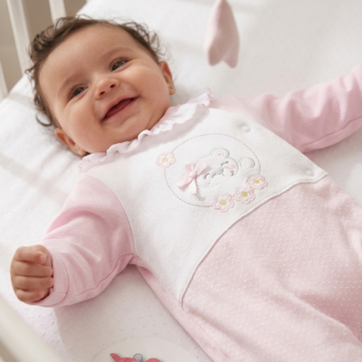 Bebé sorridente com pijama cor-de-rosa e branco com ursinho bordado numa cama branca.