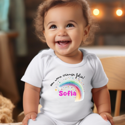 Bebé sorridente com body branco e estampa colorida de arco-íris e texto na frente sentado em cadeira de madeira