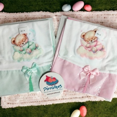 Mantinhas de bebé verdes e rosas com ursinhos bordados e laços, embaladas em plástico, sobre pano cor-de-rosa com ovos coloridos ao redor.