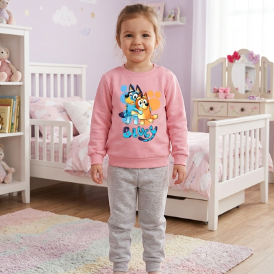 Menina sorridente com camisola cor-de-rosa com personagens Bluey e calças cinzentas em quarto decorado com móveis brancos