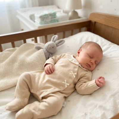 Bebé a dormir em macacão bege numa cama com cobertor e peluche coelho